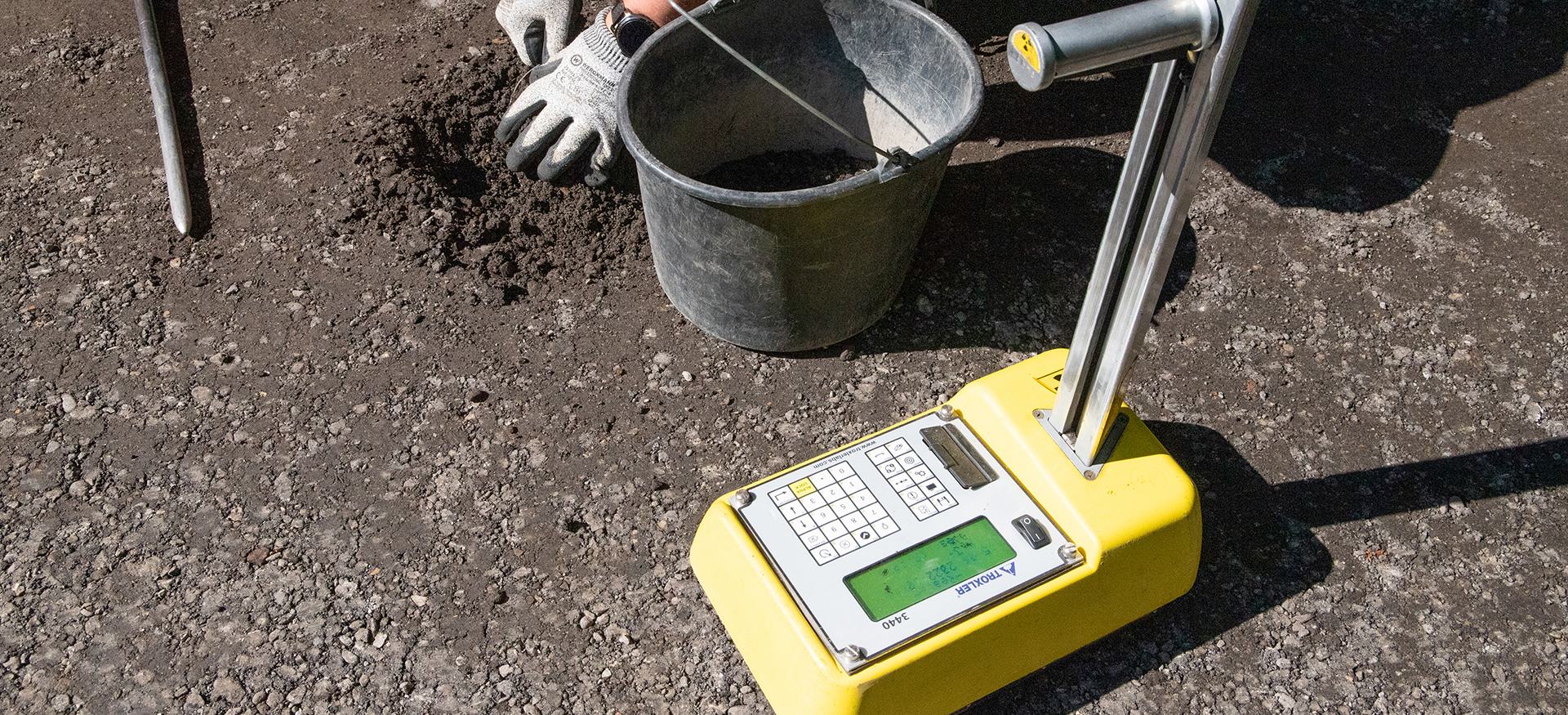 Emmer, twee handen met handschoenen in de aarde en een geel meetinstrument